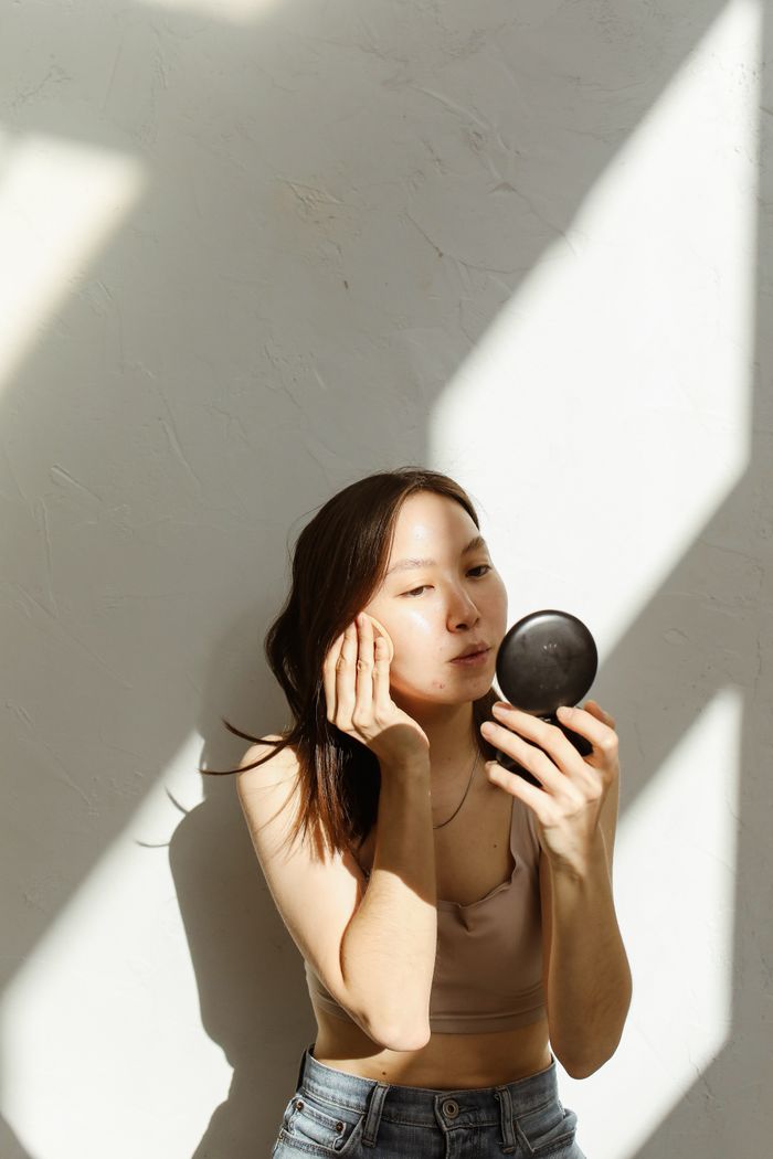 Asian woman applying face powder indoors with natural sunlight highlighting her skin.
